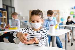 A brightly lit elementary school classroom full of children donning white protective masks and a young brunette girl in the foreground happily spraying anti-bacterial solution onto her hands.