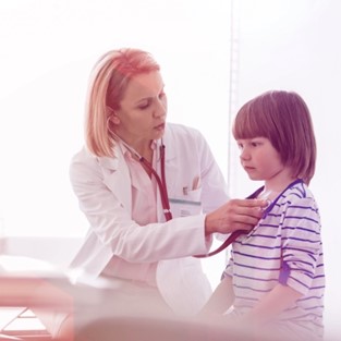 A blonde doctor in a white lab coat holding a red stethoscope to a child’s chest during an examination. The child is wearing a blue and white striped shirt.