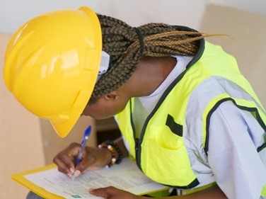 A worker with a yellow hard-hat and fluorescent yellow vest writes on a form using a blue pen.
