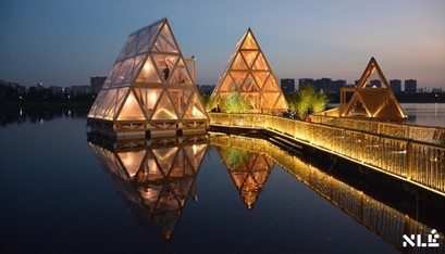 Three glowing triangular structures with triangular windows floating on the water, their images reflect in the water next to a boardwalk leading to them. Profiles of city buildings are in the background across the water.