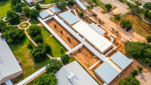 Aerial photograph of the school campus. Multiple modular buildings connected by a covered walkway on fresh dirt angled across the picture from upper left to lower right, filled out by green grass and trees.