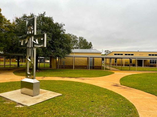 Exterior view of the Catholic School. A large metallic cross sculpture is on green grass in the lower left corner framed by a circular walkway that leads off to two buildings in the background on the right and a line of trees to the left.