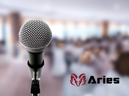 Blurred background photo of a conference room. At the left is an in focus, close-up of a microphone and on the lower right is a ram’s head logo in red and in black letters the word “Aries.”