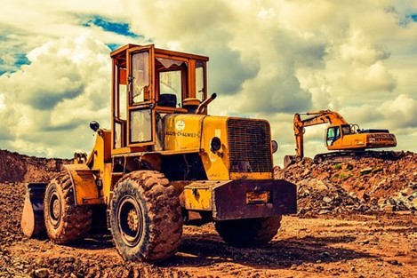 A photograph of an excavator and a bulldozer, the excavator is further away and up on a large mound of dirt. They both sit ready to work on a red dirt site with a sky full of big clouds above and behind them.
