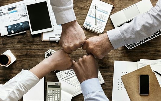 Above a wooden desk strewn with work elements (calculators, phones, tablets, newspapers, pens, and laptops) the hands of four people meet in a friendly fist bump.