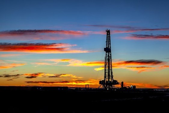 A setting sun paints the sky red, purple, and orange and leaves a large oil drill in the shadows surrounded by darkness.