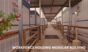 : A photograph of a covered wooden boardwalk connecting several aligned blue and white modular units both on the left and right with the Aries’ logo and phone number printed on a sign. The phrase, “WORKFORCE HOUSING MODULAR BUILDING” is in white text on the bottom right.