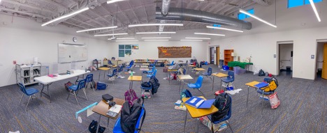 The inside of a clean, well-lit classroom with no people inside and focusing on the desks and chairs with a lot of space between them and books and backpacks populating each desk.
