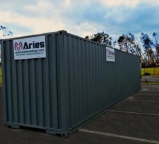 A photograph of a steel matte gray mobile storage container resting in a parking lot with a cloudy blue sky and a line of trees in the background.