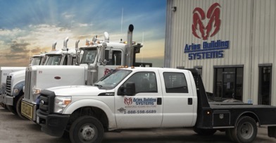 Photograph of four various types of white trucks parked in a row in front of a building with a sunrise behind them. “Aries Building Systems” is on the truck in front and on the building with the red ram logo as well.