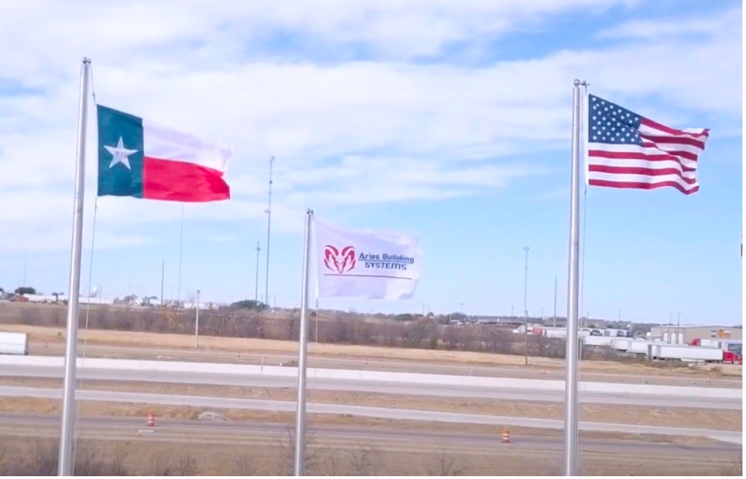A photograph of the Texas state flag, Aries’ flag, and the American flag flapping proudly in the wind against a cloudy sky outside the Aries’ modular building facility in Troy, Texas.