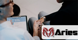 Photo of two people engaging in a handshake. The person on the left is an employer wearing white professional dress clothing and holding a clipboard with a resume. The person on the right is the jobseeker wearing black professional dress clothing. Aries’ red ram logo is on a white rectangular background on the bottom right corner of the photograph.