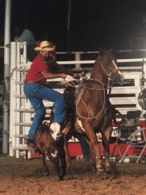 VisionAries Dee Stephens in his cowboy hat and attire dismounting from horse to rope a calf that is in the foreground