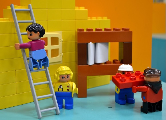 one Lego person up on a ladder while another assists them below. Two other Lego figures help each other bring a red brick to the yellow wall being constructed