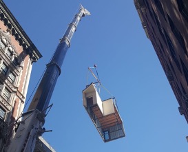 a modular home section being lifted by a crane high above and between buildings.