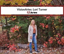 VisionAries, Lori Turner, posing in front of beautiful trees filled with fall colored leaves. At the top is a white banner with a thick red border that reads, “VisionAries: Lori Turner” and the red ram Aries’ logo under it.