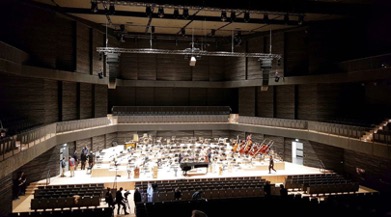 : The interior of the Isarphilharmonie concert hall with an orchestra practicing and lights on the stage. There is no audience yet.