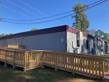 A gray modular building with dark red trim and air conditioning units on the exterior. The building is marked “E6” and sits behind a wooden ramp leading up to its entrance.