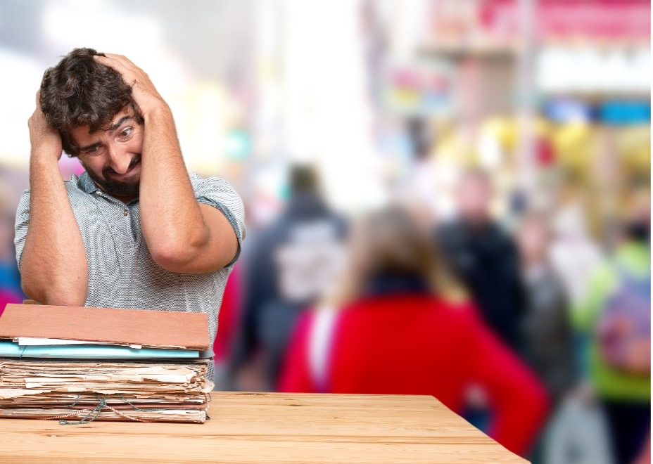 In a blurred background school setting, a person with their head in their hands conveying a sense of stress or overwhelm, representing a teacher or administrator with space limitations standing behind a table overflowing with folders.