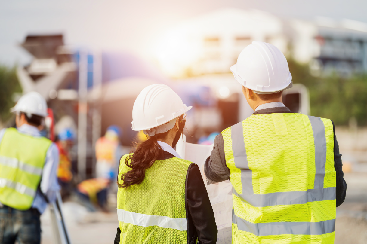 The backs of two construction workers discussing plans at an active construction site.