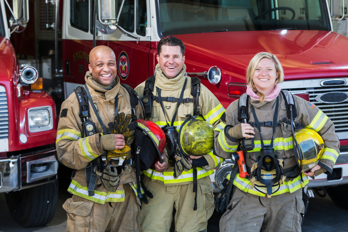 Fire fighters smiling in uniform in front of fire trucks.