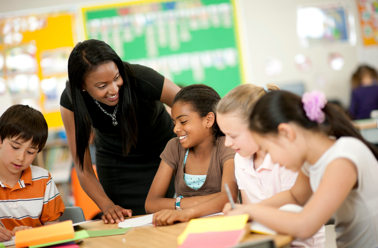 A smiling teacher helping students at their desks inside of a classroom.