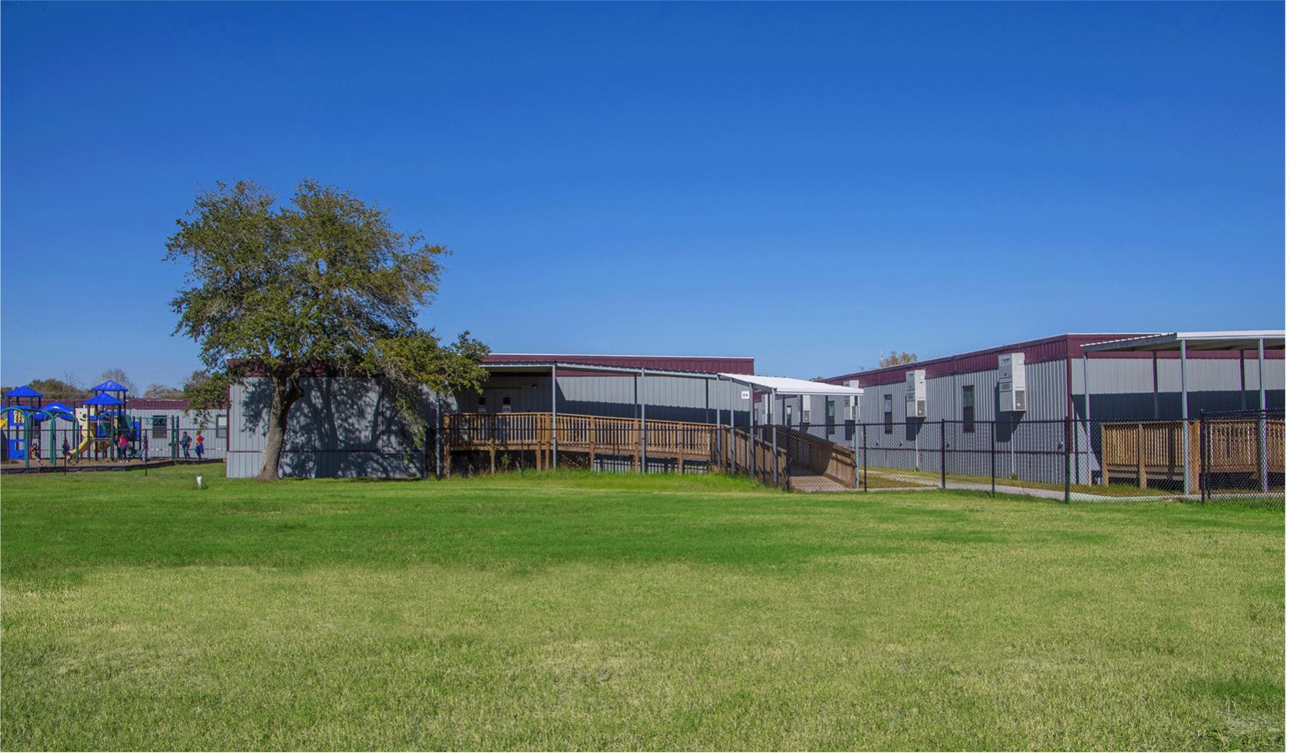 Several Aries portable classrooms on a green lawn during a sunny day. They have wooden ramps and are located to near a playground.