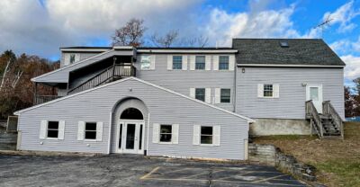 A photo of the beautiful Aries Windham, NH ofice. There is a smaller house-like building in front of a larger building with a staircase leading to the upper levels. Both buildings are paneled in light gray wood with white shudders on all of the windows.