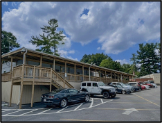 Modular building with deck and patio covering, wooden steps down to a parking lot, and parked cars on a sunny day.