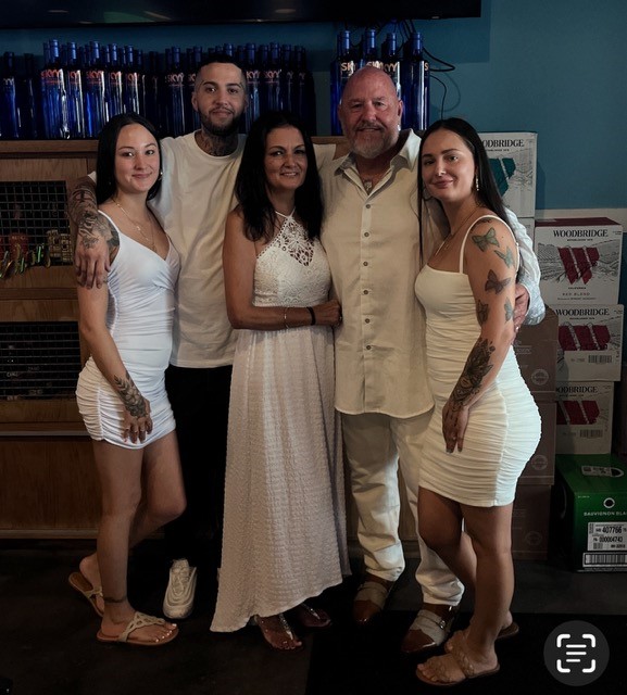 A photo of Ash Mote, his wife, two daughters, and son all smiling in formal white attire.