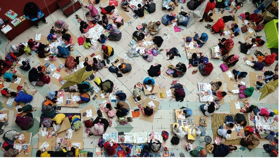 Overhead view of a crowded classroom or event space filled with students working on activities at small tables and on the floor.