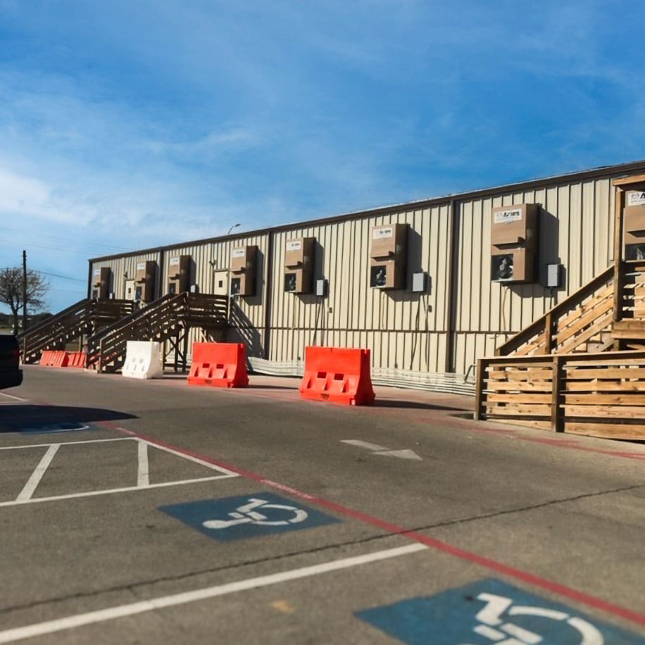 A row of GSA mobile office units with wooden staircases and orange and white safety barriers in the parking lot, equipped with external air conditioning systems.