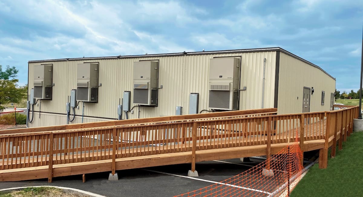 A beige modular building with four large air conditioning units mounted on its side is shown. In front of the building, there is a wooden ramp with railings, leading to an entrance on the right side.