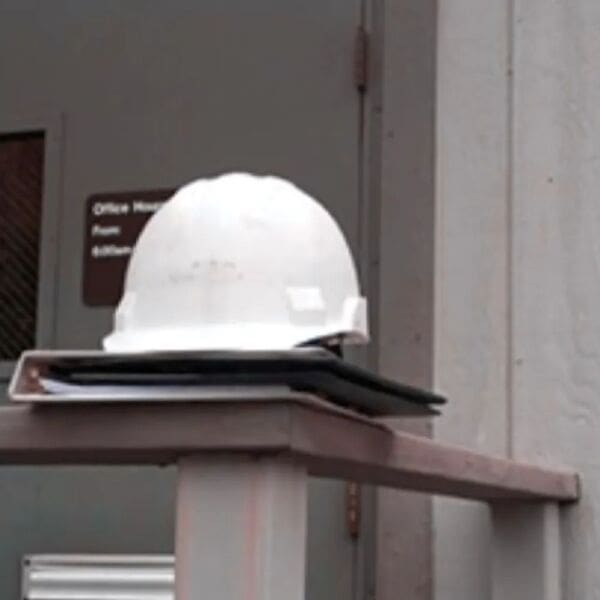 A construction worker’s hard hat resting on a binder balanced on a railing from the outside structure of a jobsite trailer.