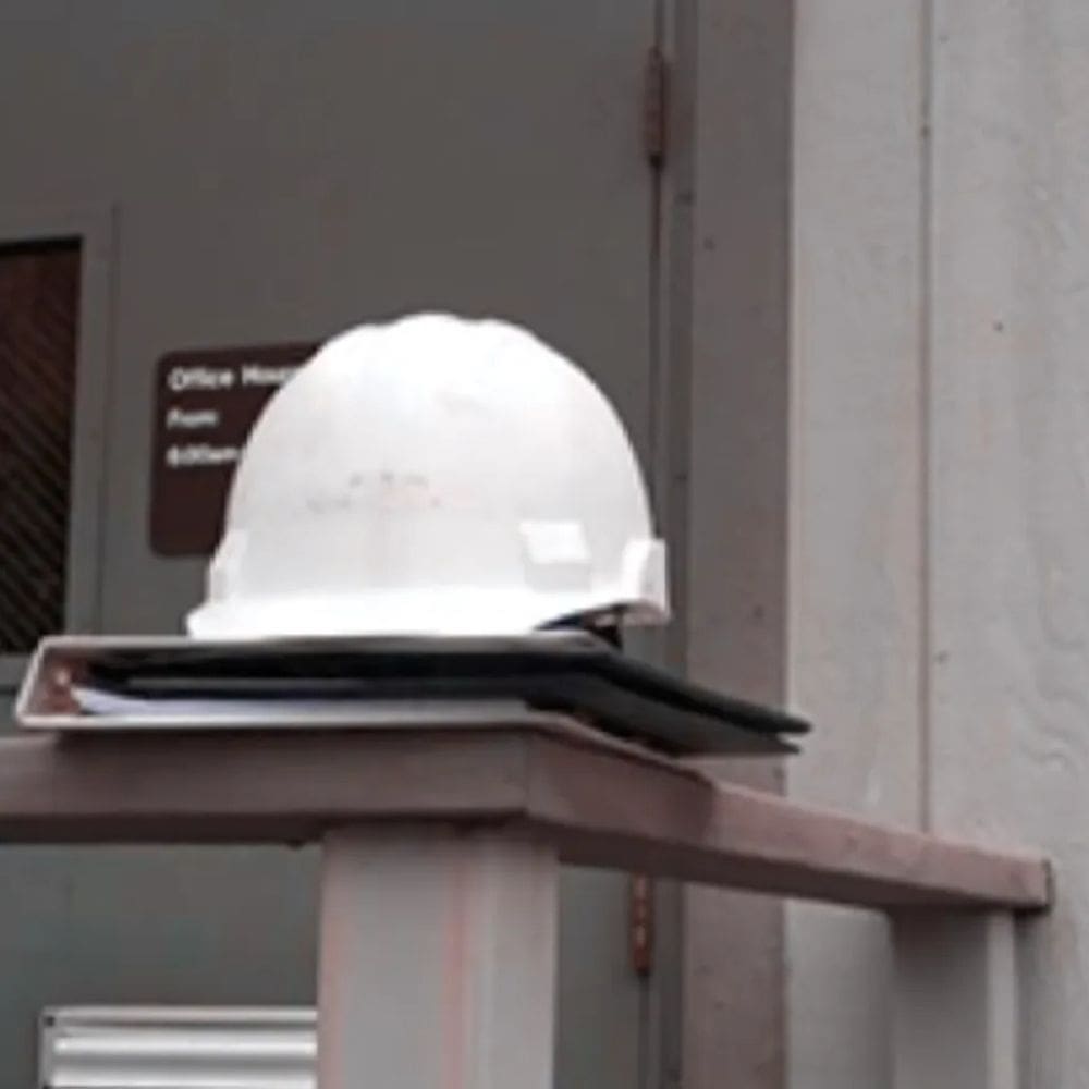 A construction worker’s hard hat resting on a binder balanced on a railing from the outside structure of a jobsite trailer.